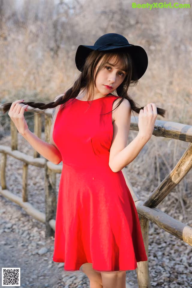 A woman in a red dress and black hat leaning against a fence.